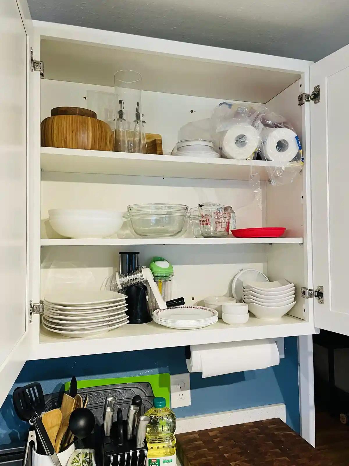 An open white kitchen cabinet filled with neatly stacked plates, bowls, measuring cups, wooden bowls, and paper towels, with…