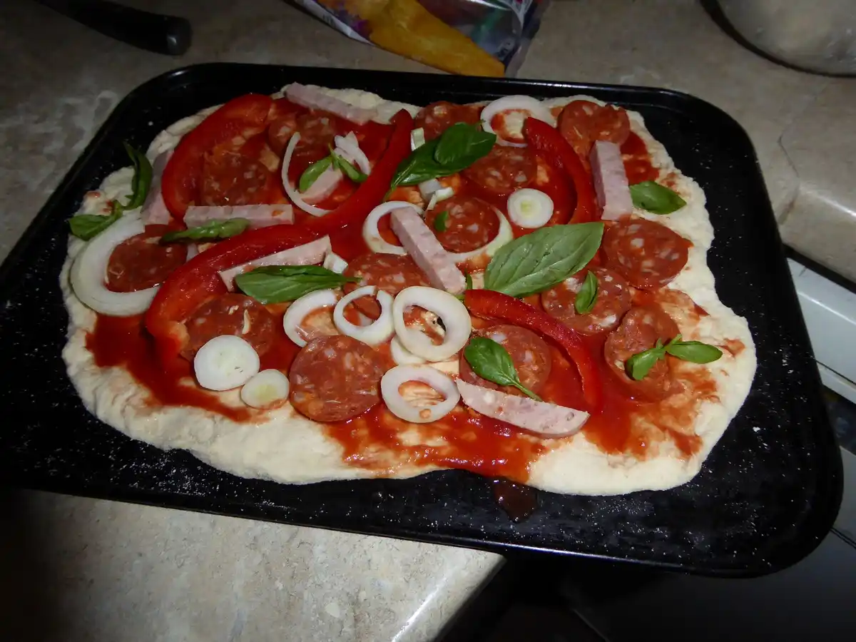 An uncooked pizza on a black baking tray, covered in tomato sauce, pepperoni, sliced red bell peppers, onion rings, basil…