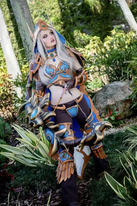 A person in a detailed fantasy cosplay of a female archer with blue and gold armor, white hair, and a large ornate bow…