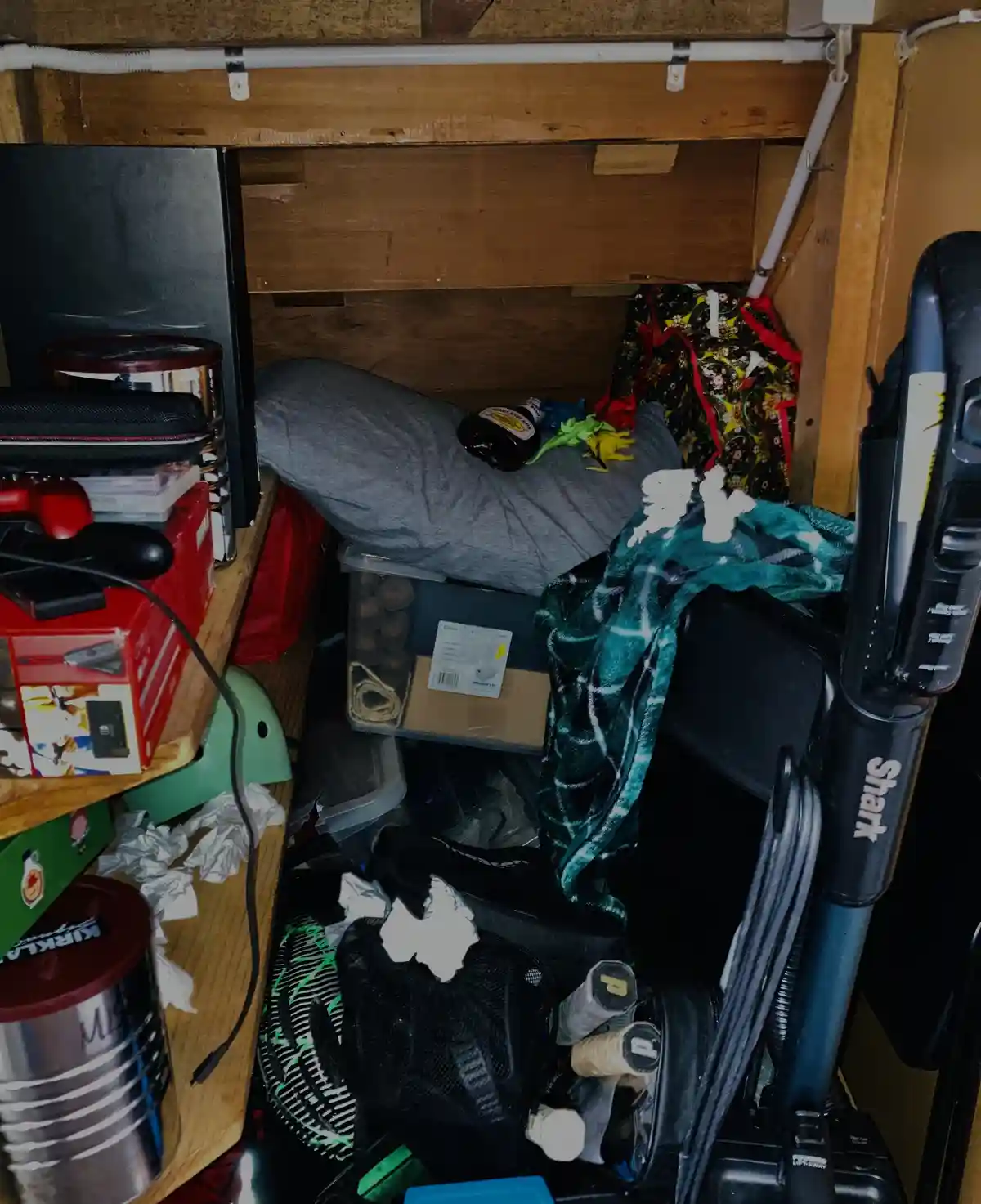 A cluttered, cramped closet filled with various items including a Shark vacuum, storage bins, a pillow, a bottle, and…