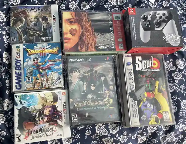 A collection of video game cases and a controller box laid out on a floral-patterned fabric, including titles like…