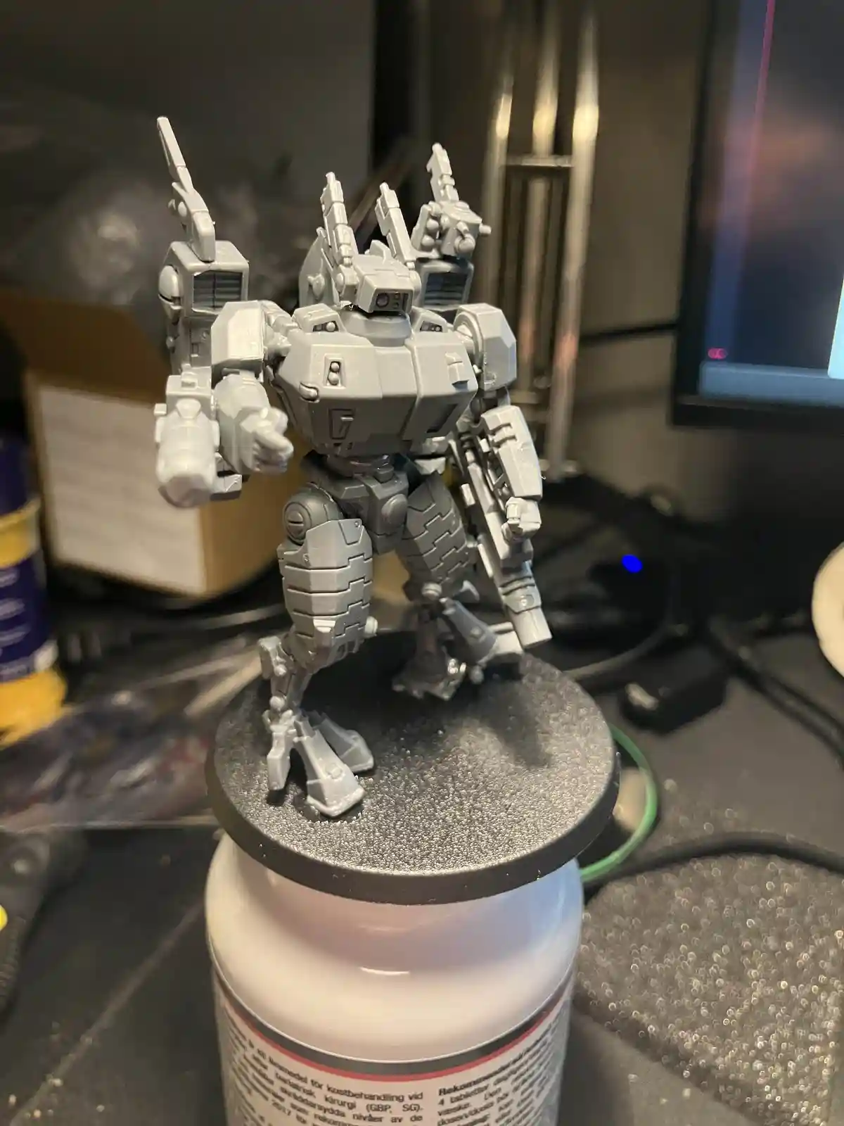 A gray, unpainted, heavily armed robot model stands on a black circular base, which is placed atop a white pill bottle. The…