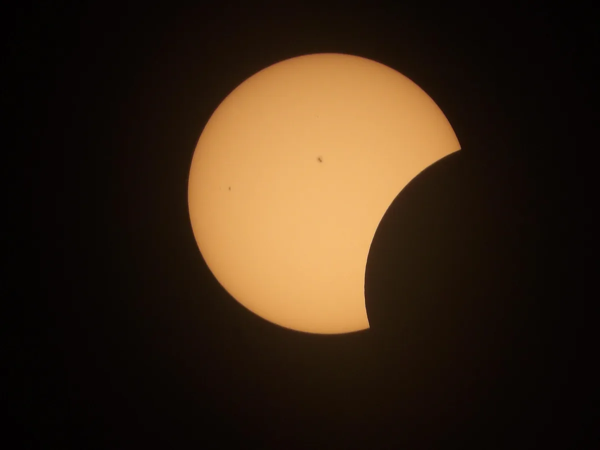 A partial solar eclipse shows the sun as a bright orange crescent against a black sky, with small dark spots visible on its…