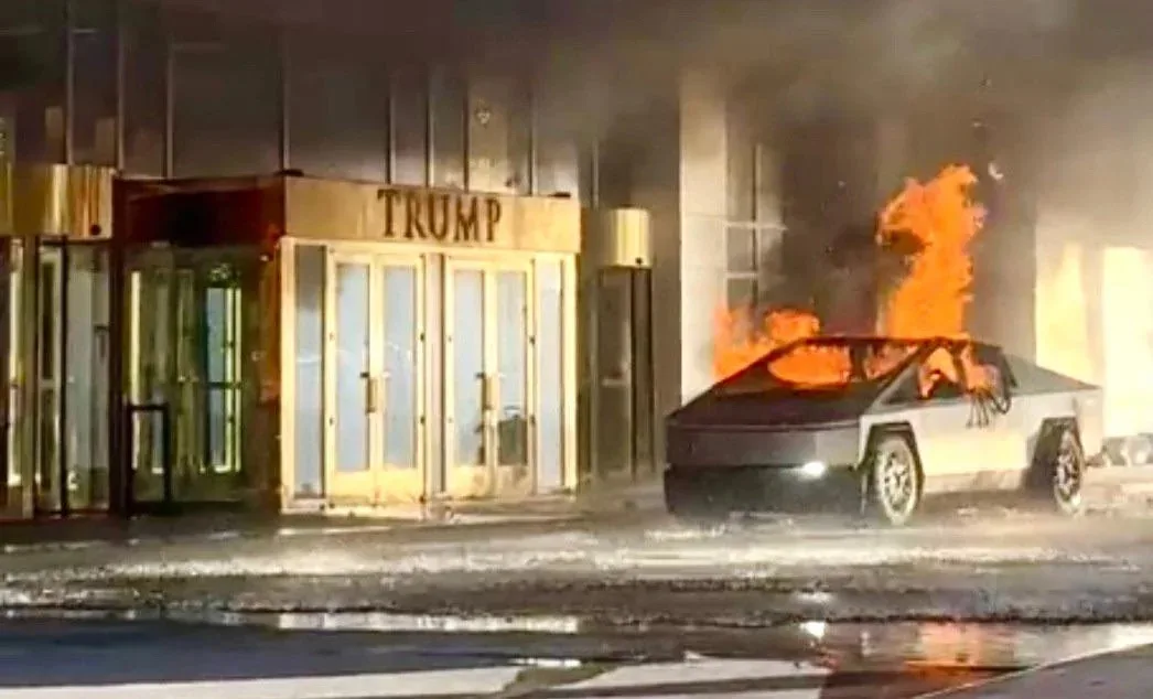 A white Tesla Cybertruck is engulfed in flames on a street at night, with thick black smoke rising. Behind it, a building…
