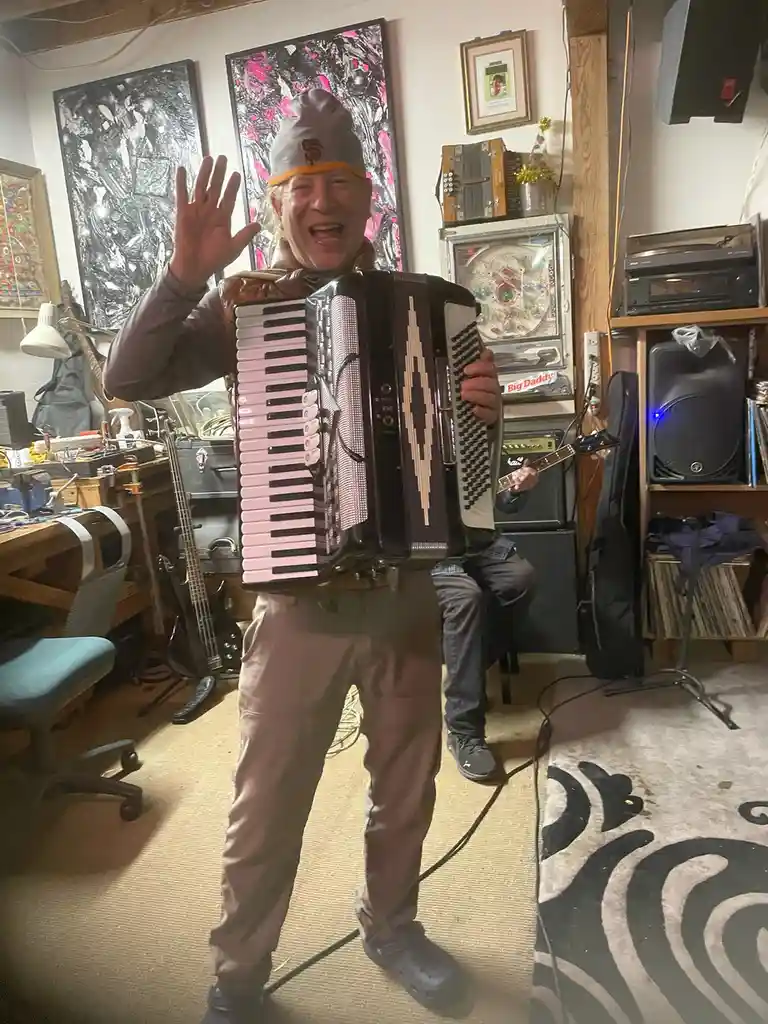 A cheerful older man in a grey beanie and brown pants stands in a cluttered music room, enthusiastically playing a large…