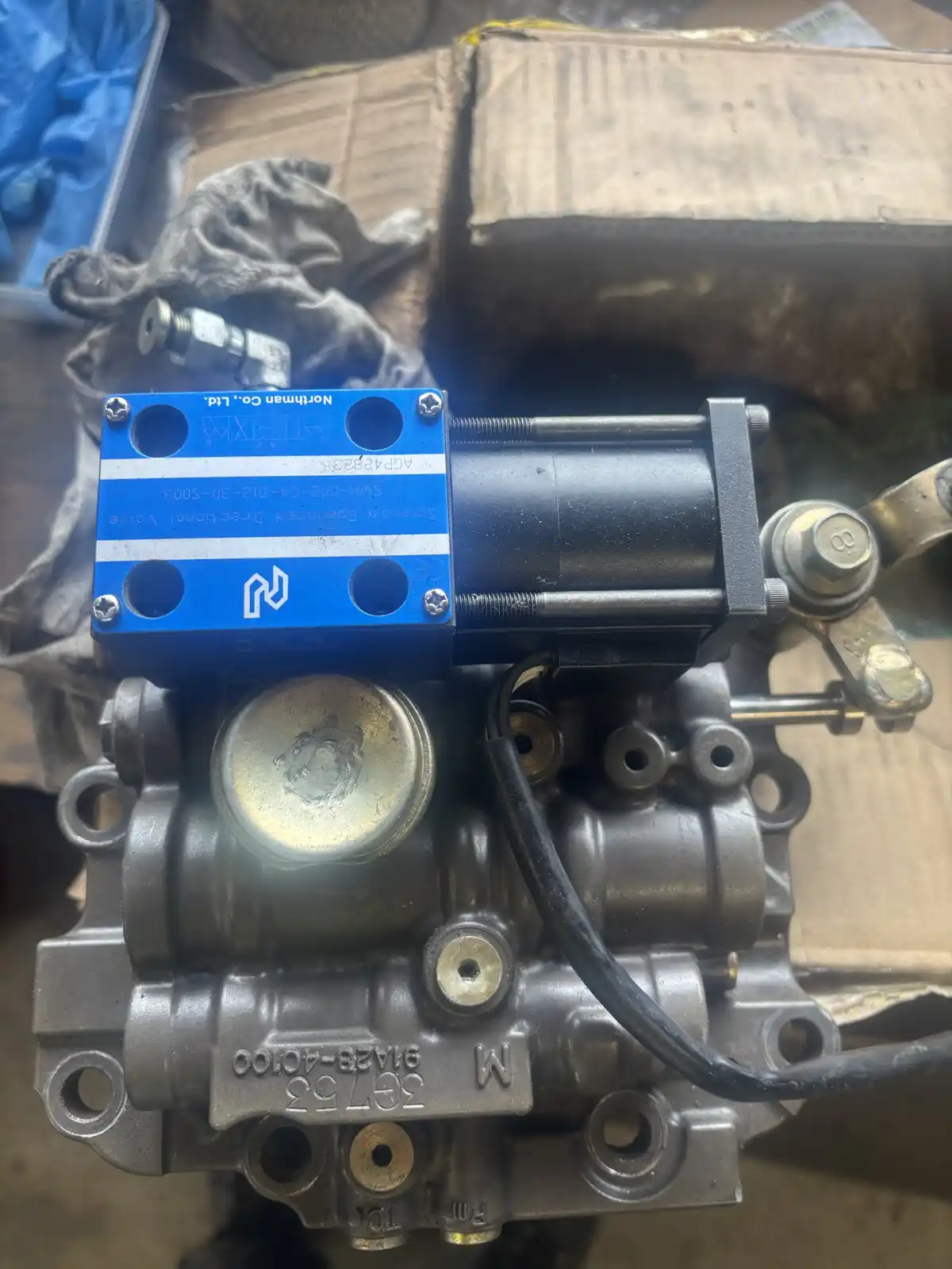 A close-up photo shows a mechanical component, likely a transmission valve body, with a blue solenoid valve mounted on top…