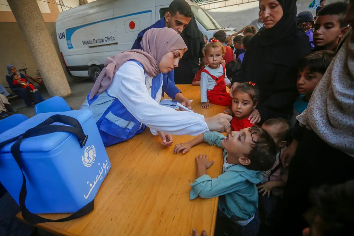 A UNICEF worker in a blue vest examines a young boy's mouth at a mobile dental clinic, surrounded by children and adults. A…