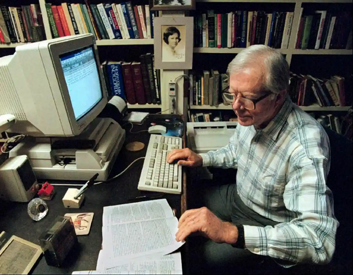 An older man with glasses sits at a cluttered desk surrounded by bookshelves, typing on a beige computer keyboard while…