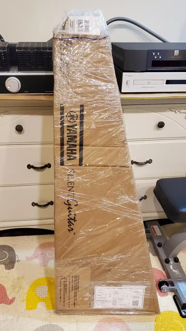 A tall, narrow, brown cardboard box wrapped in clear plastic, labeled 'YAMAHA SILENT Guitar™', stands upright on a colorful…