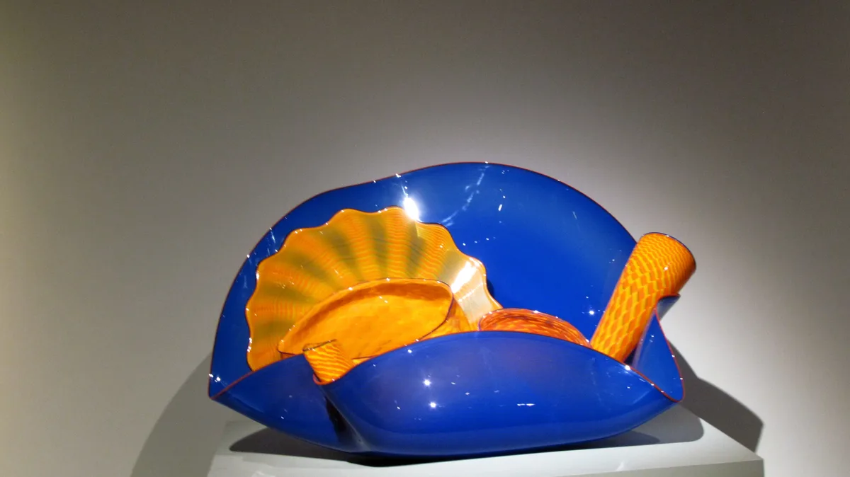 a large, glossy blue glass sculpture shaped like a chair with orange and yellow wavy patterns, displayed on a white pedestal…