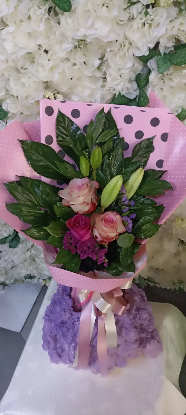 a bouquet of pink and magenta roses, green lilies, and purple filler flowers is wrapped in pink polka dot paper and tied…