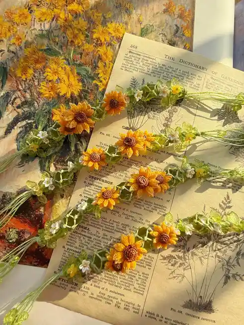 A handmade beaded sunflower crown with green leaves and small white flowers rests on an open vintage dictionary page titled…