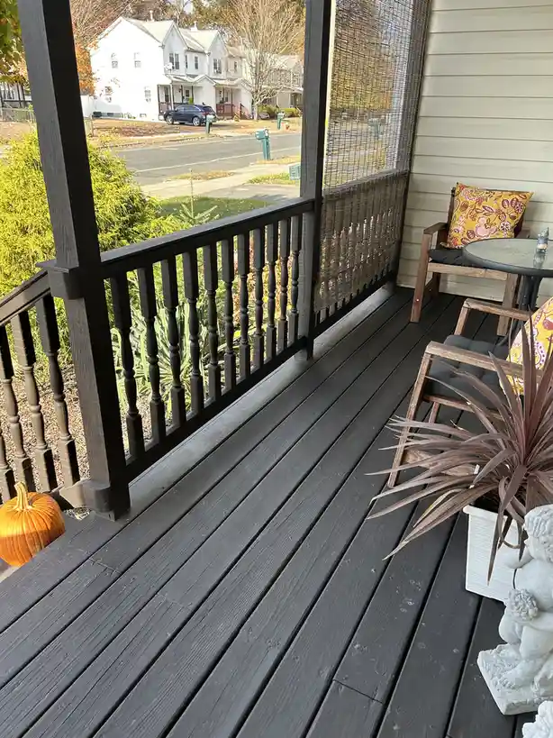 A photo of a dark-stained wooden porch with black railings, featuring a small orange pumpkin, a potted plant, and outdoor…