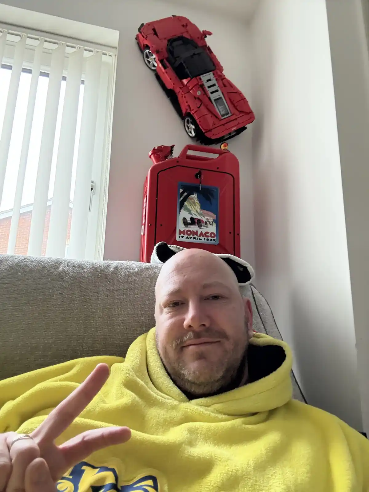A bald man in a yellow hoodie sits on a couch making a peace sign, with a red LEGO Ferrari model mounted on the wall above…