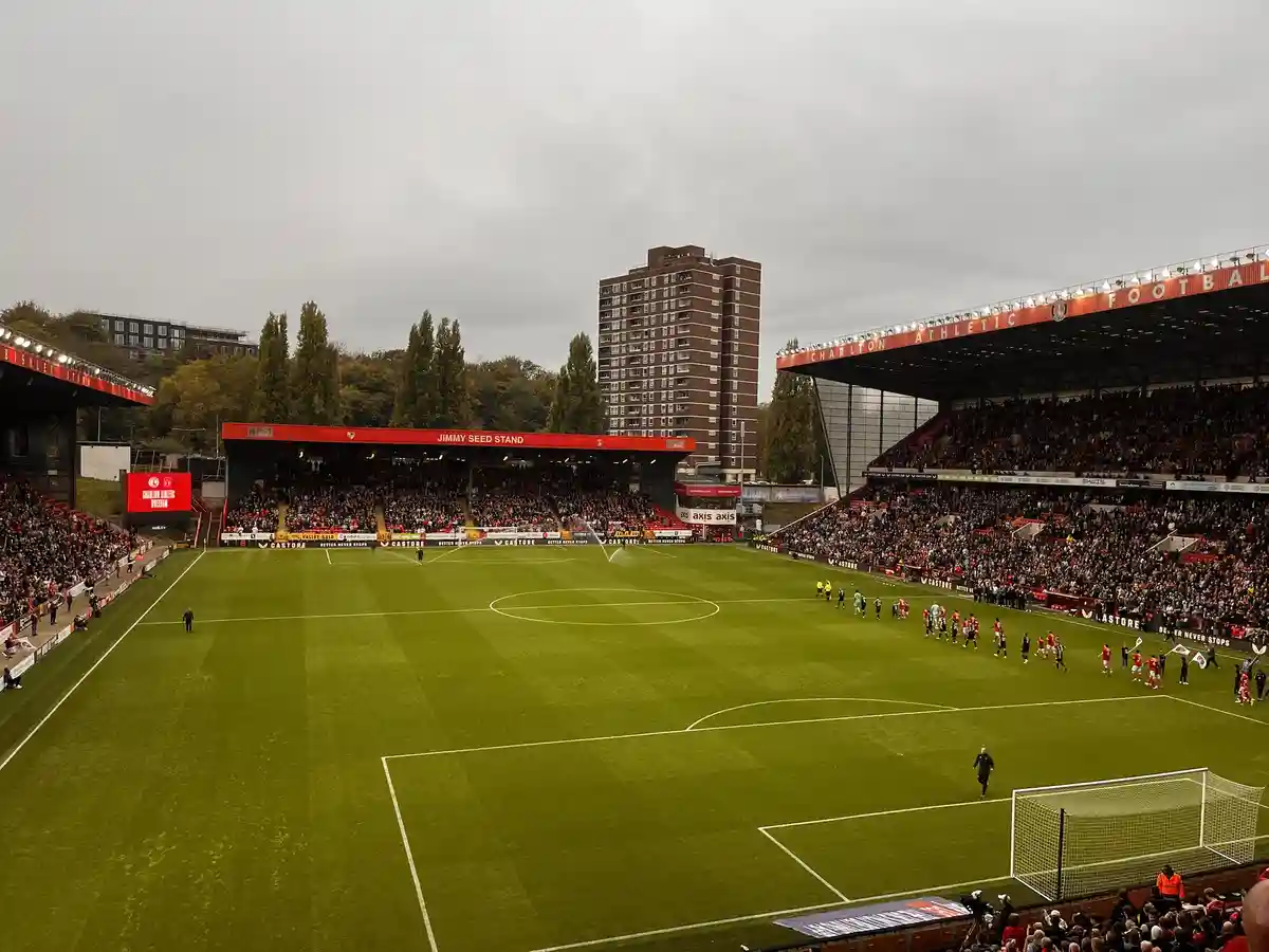 A wide shot of a packed football stadium with a green pitch, red seats, and a large building visible behind the stands. The…
