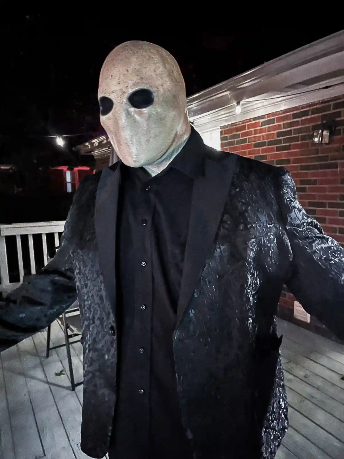 A person wearing a blank white mask with black eye holes and a black tuxedo jacket covered in crinkled black plastic stands…