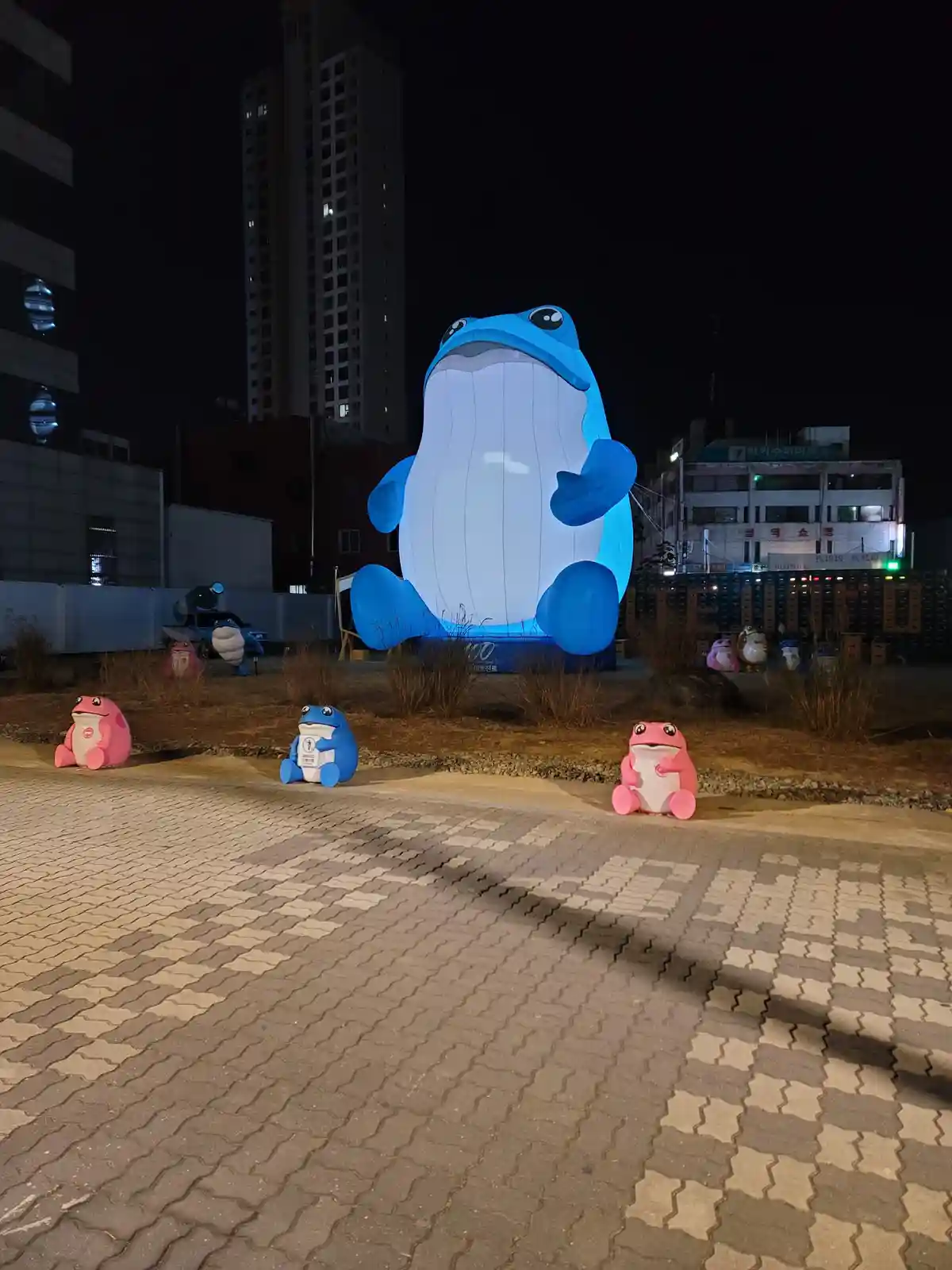 A nighttime photo shows a large, illuminated blue inflatable whale-like creature sitting in an urban area, with three…