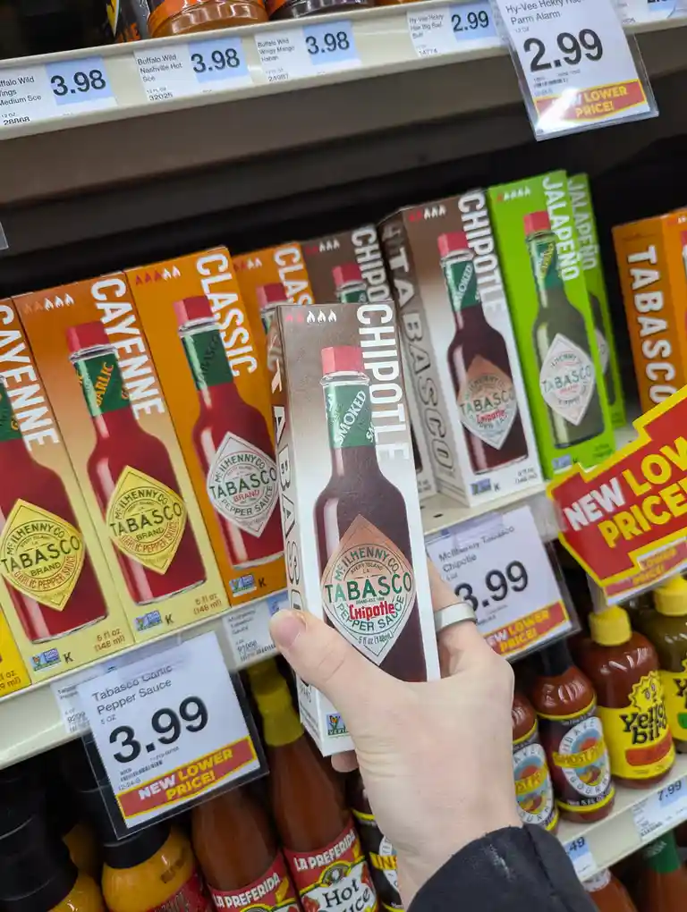 A hand holds a box of Tabasco Smoked Chipotle Pepper Sauce in a grocery store aisle, surrounded by other Tabasco varieties…