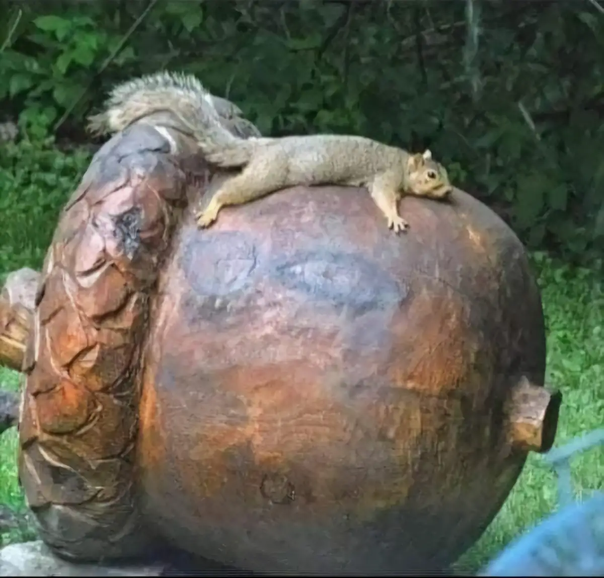 A small squirrel is lying stretched out on top of a large, weathered wooden sculpture resembling a human head, with green…