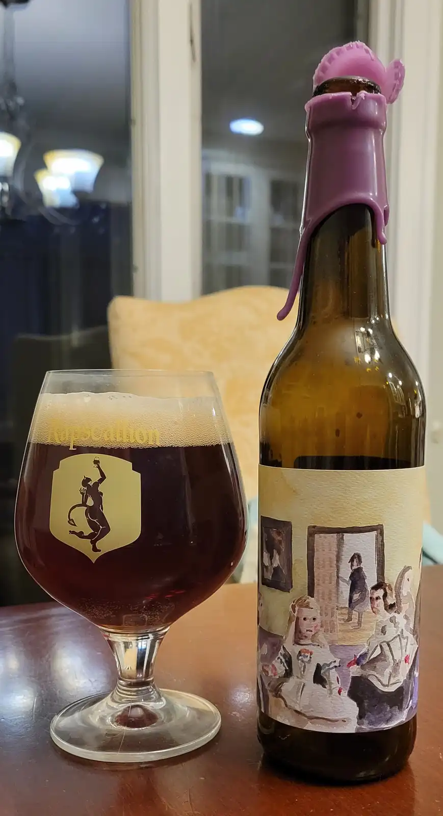 A dark beer in a glass labeled 'Rapscallion' sits next to its bottle, which has a purple silicone bottle cap and a label…