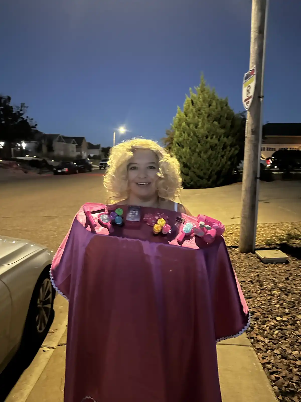 A woman with a blonde wig stands outdoors at dusk, smiling while wearing a purple cape draped over her shoulders. The cape's…