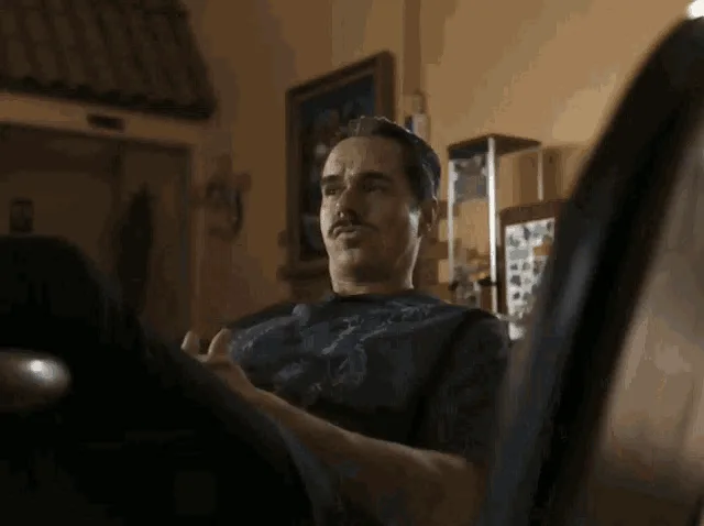 A six-panel image sequence shows a man with a mustache in a dimly lit room. In the first three panels, he sits in a chair…