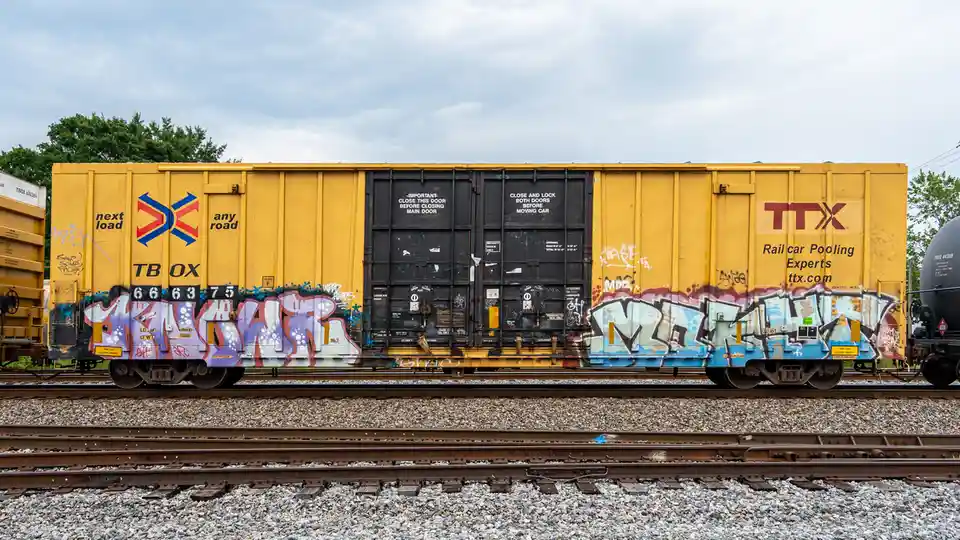 A yellow TTX railcar with graffiti on its sides sits on train tracks under a cloudy sky. The black doors have white safety…