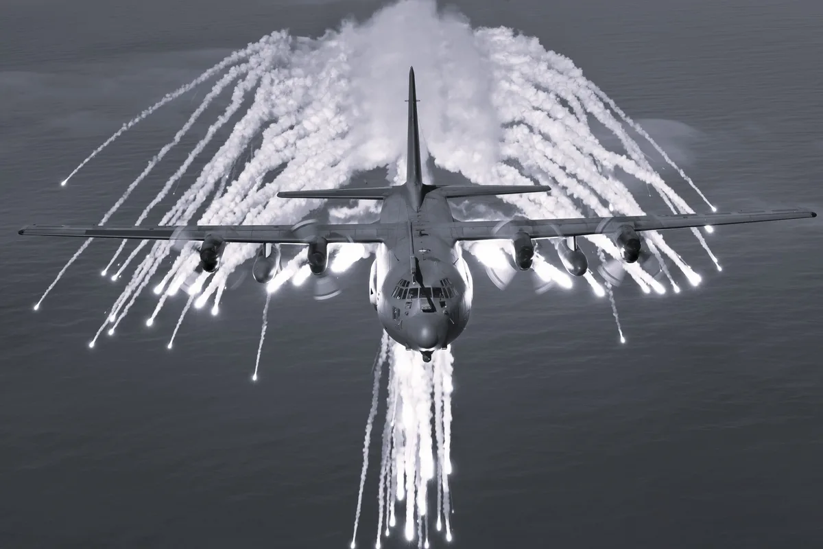 A black-and-white photo shows a military transport aircraft flying over water, deploying flares from its wings and fuselage…