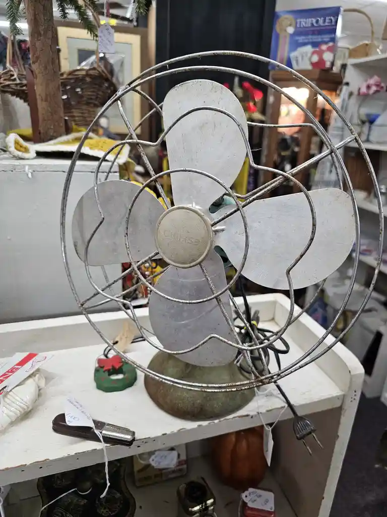 A vintage, rusty metal fan with four blades and a wire cage sits on a cluttered thrift store shelf, surrounded by other…