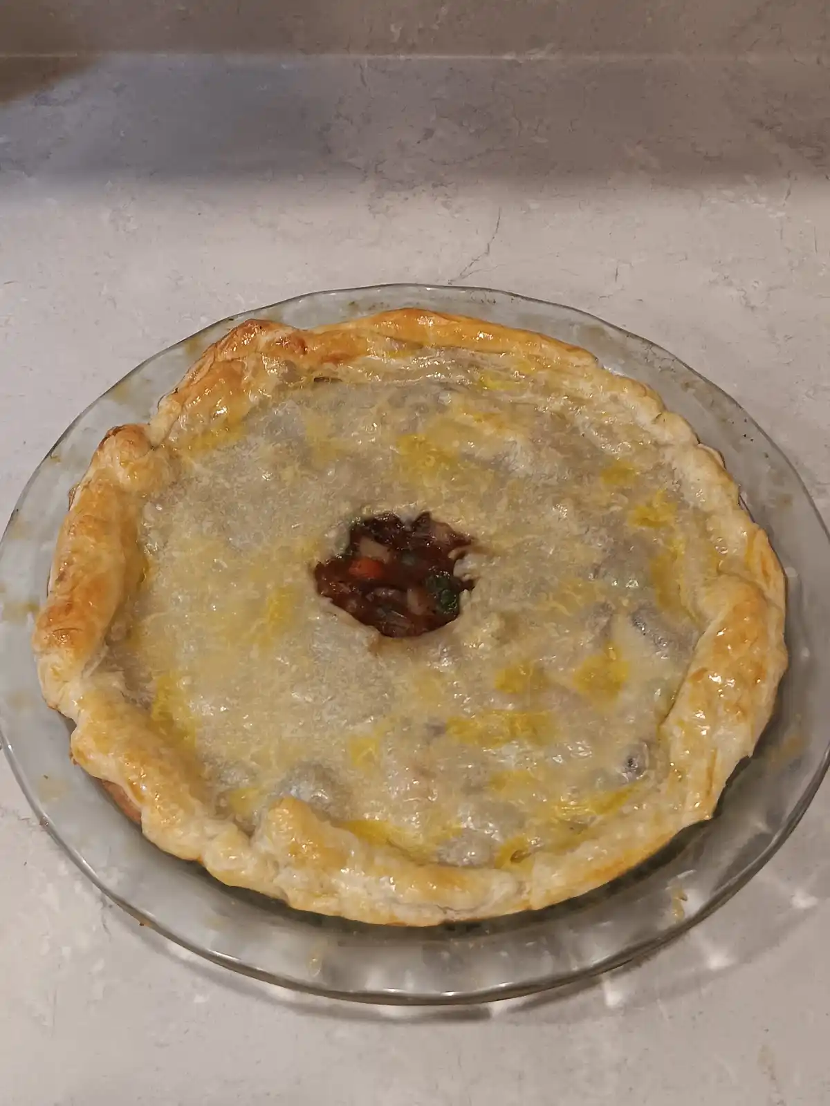A golden-brown baked pie sits in a clear glass dish on a light-colored countertop, its crust slightly puffed and glazed. A…