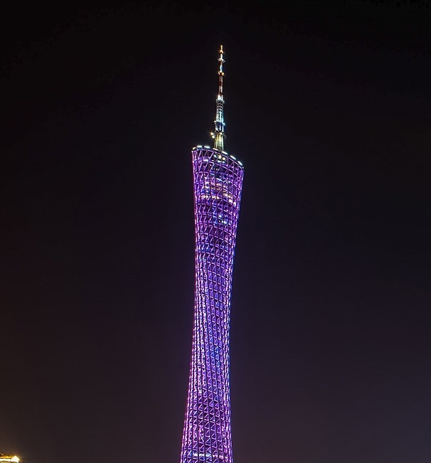 A tall, slender tower with a distinctive curved, lattice-like structure is illuminated in purple and blue lights against a…
