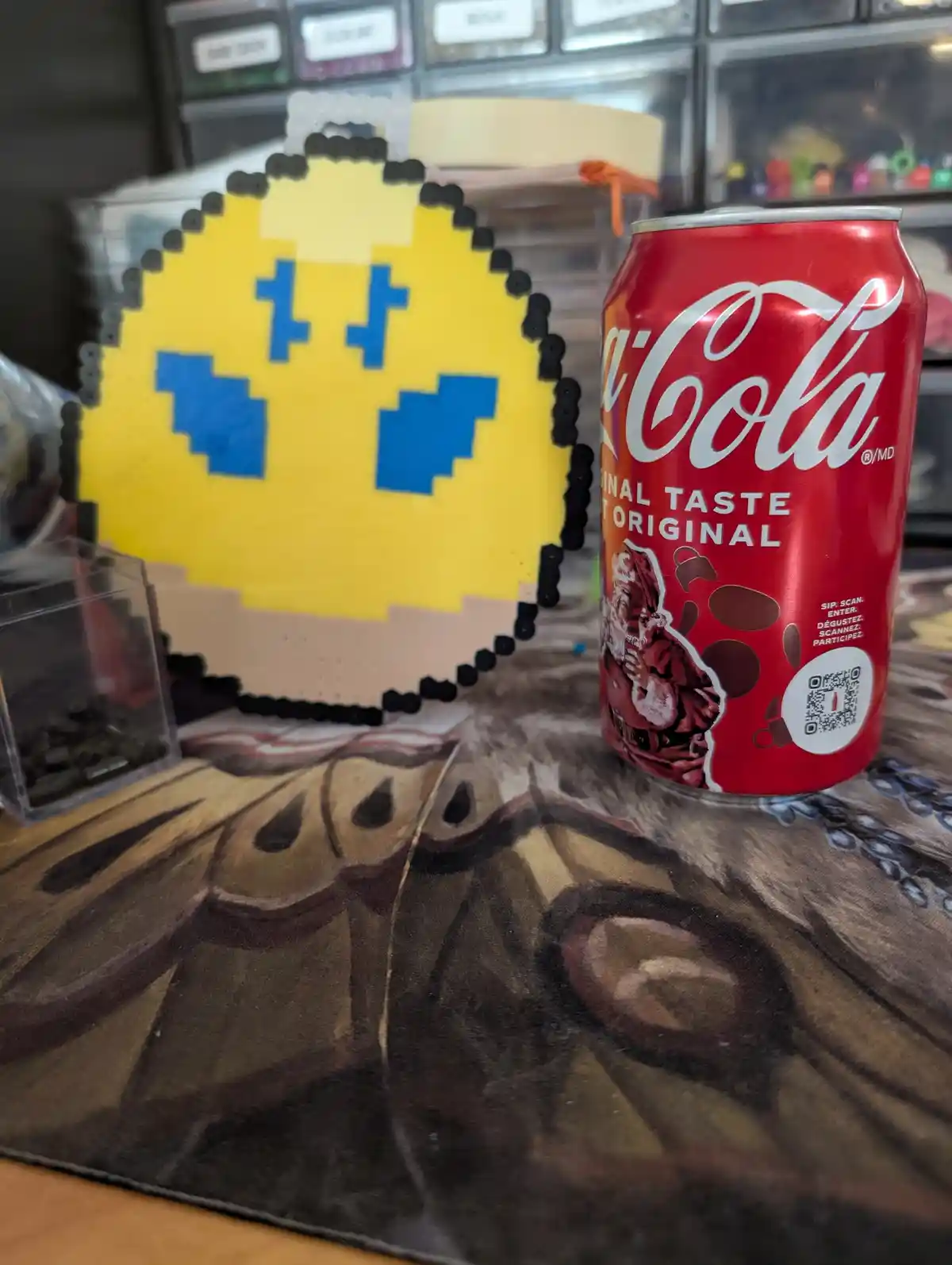 A red Coca-Cola can sits next to a handmade pixel art Pac-Man figure made of beads. The can has a Santa Claus design and a…