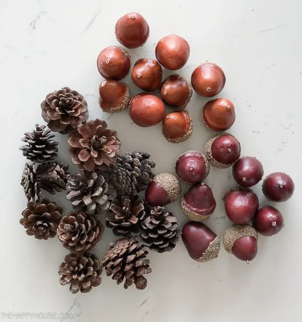 An overhead shot displays pinecones and acorns arranged on a white marble surface to form the shape of a face, with…