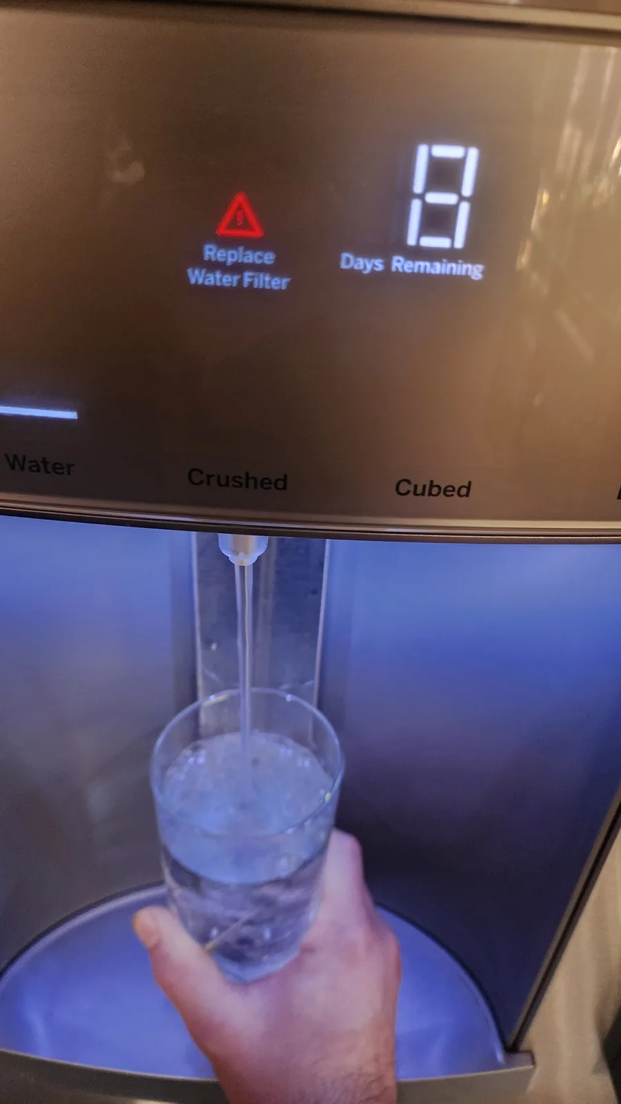 A close-up photo shows a hand holding a glass under a water dispenser, while the machine's digital display shows a red…