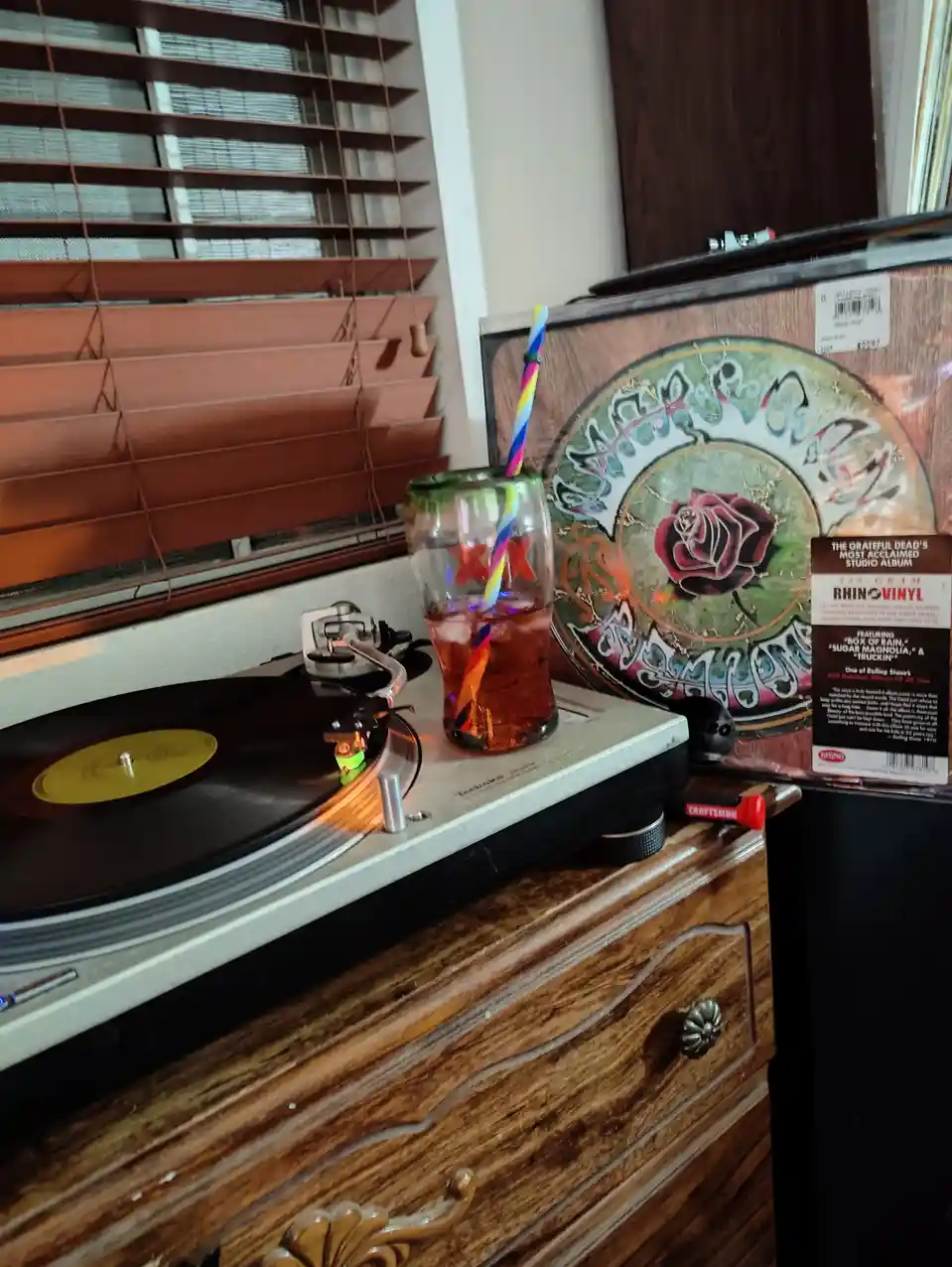 A Technics turntable spins a vinyl record on a wooden dresser, next to a glass of iced tea with a colorful straw. Behind it…