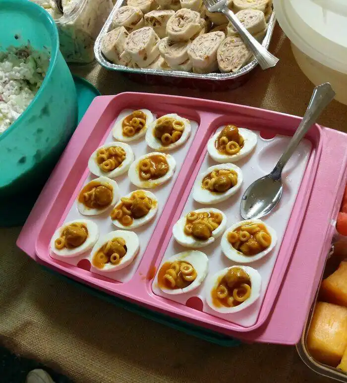 A pink egg mold tray holds deviled eggs filled with macaroni and cheese, with a spoon resting in the tray. Other party foods…