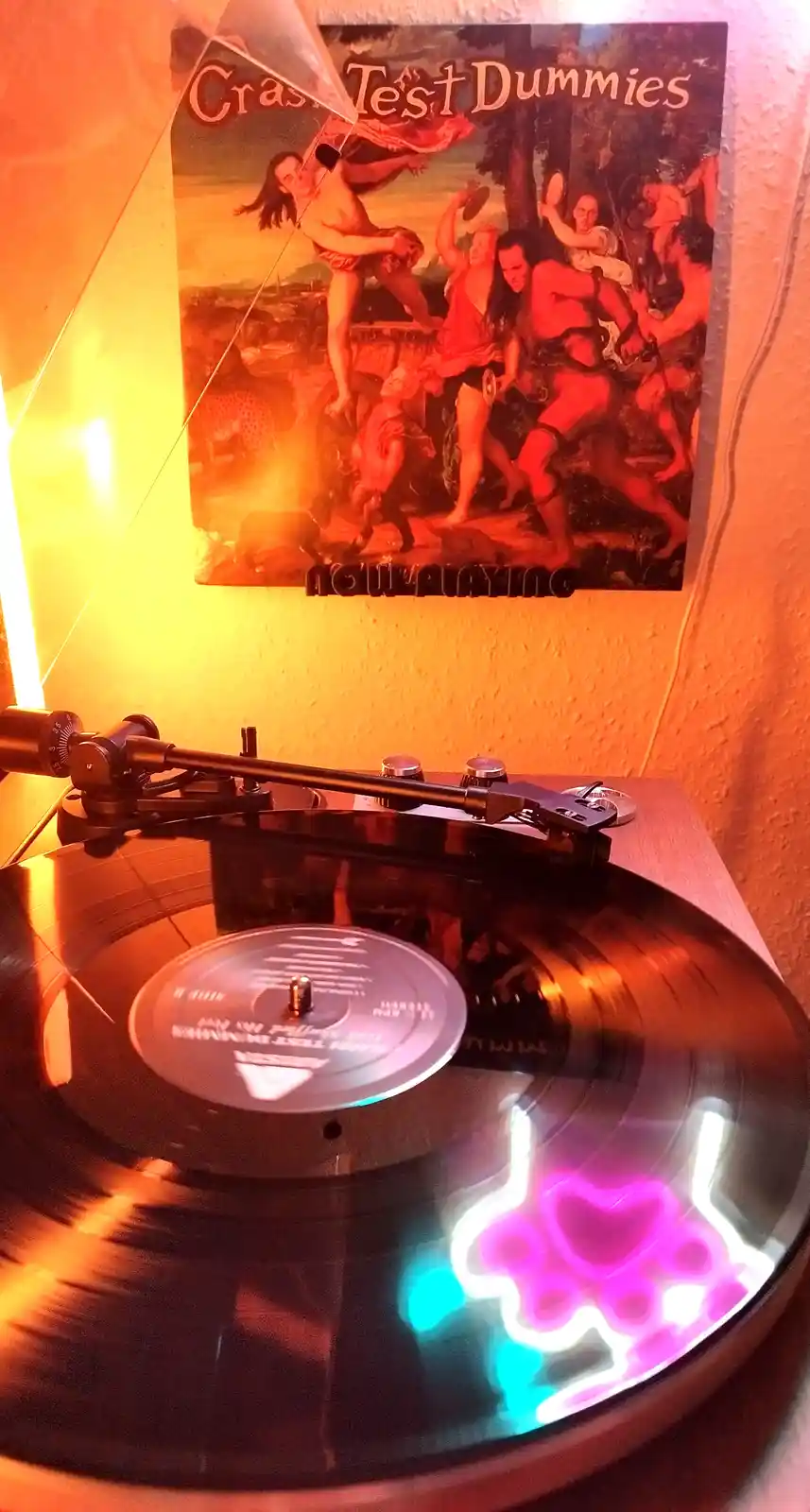A vinyl record spins on a turntable with a glowing paw print design on its surface, beneath a poster for the band Crash Test…