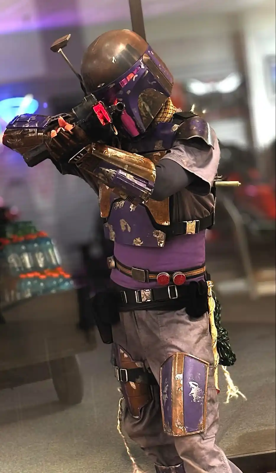 A person in a detailed, weathered Boba Fett costume, complete with helmet, armor, and blaster rifle, is standing indoors in…