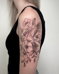 A person with blonde hair and a black tank top shows off a detailed black-and-white tattoo on their left upper arm and…
