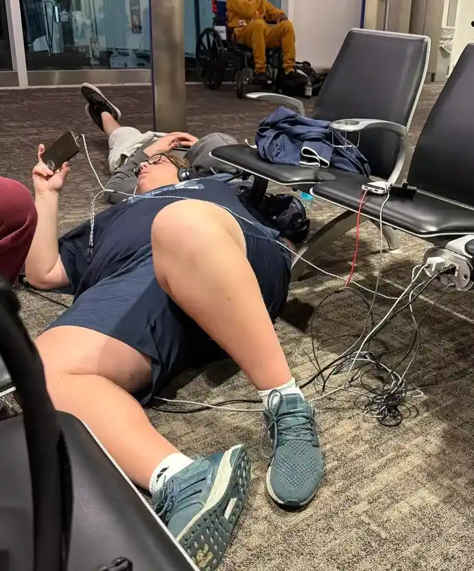 A person is lying on the carpeted floor of what appears to be an airport or waiting area, surrounded by chairs. They are…