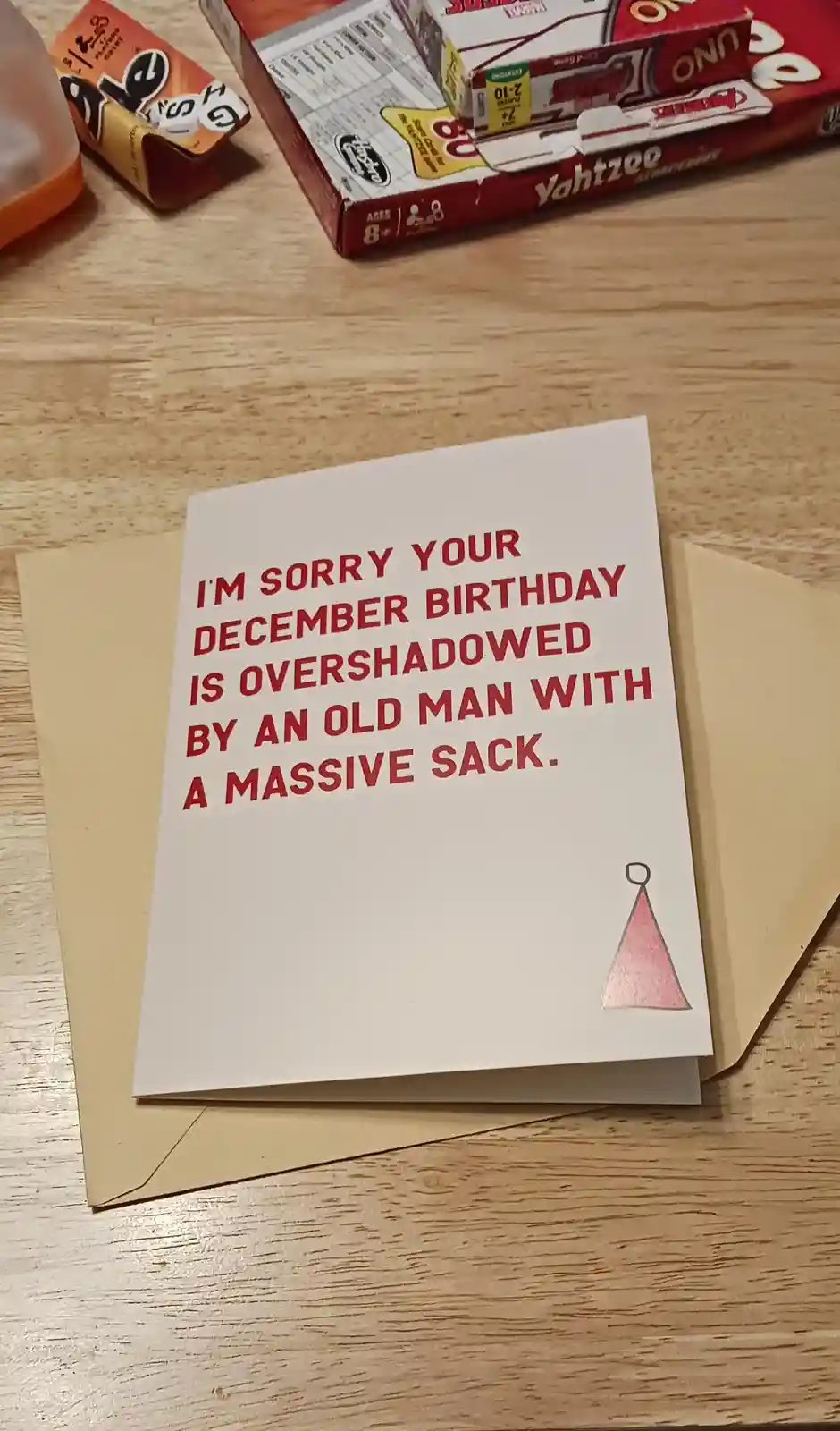 A white birthday card with red text sits on a brown envelope on a wooden table, surrounded by board games like Yahtzee and…