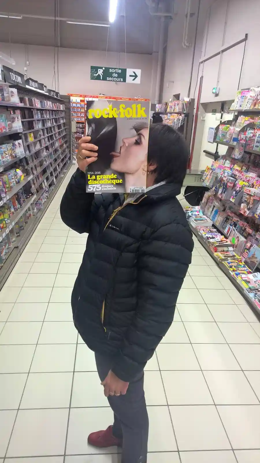 A person in a black puffer jacket stands in a store aisle holding a 'rock&folk' magazine in front of their face, obscuring…