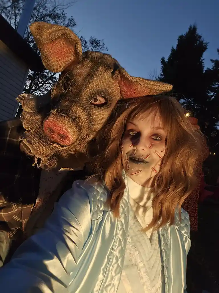 A person in a light blue satin robe with zombie makeup featuring red stitches poses for a selfie outdoors at dusk, with…