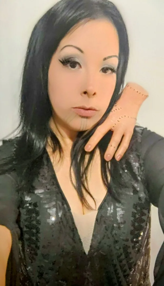 A woman with long black hair and heavy makeup poses with her hand near her face, but her hand is replaced with a mannequin…
