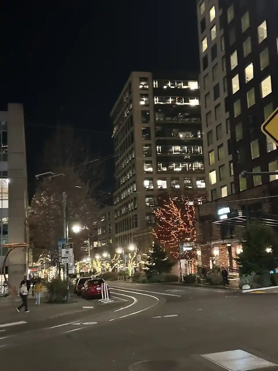 A nighttime city street scene with illuminated buildings, decorated trees with lights, and pedestrians. A red car is parked…