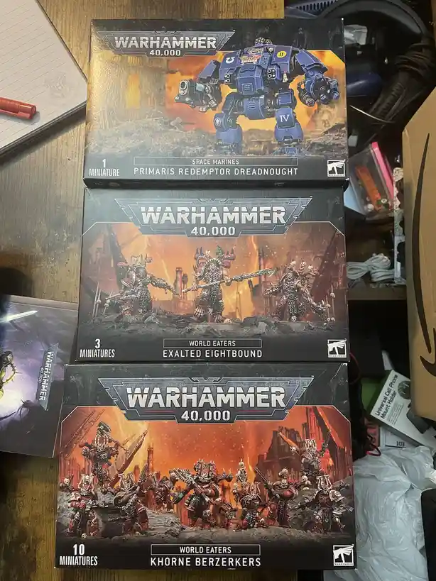 Three Warhammer 40,000 miniatures boxes are stacked vertically on a cluttered desk. The top box contains one Primaris…