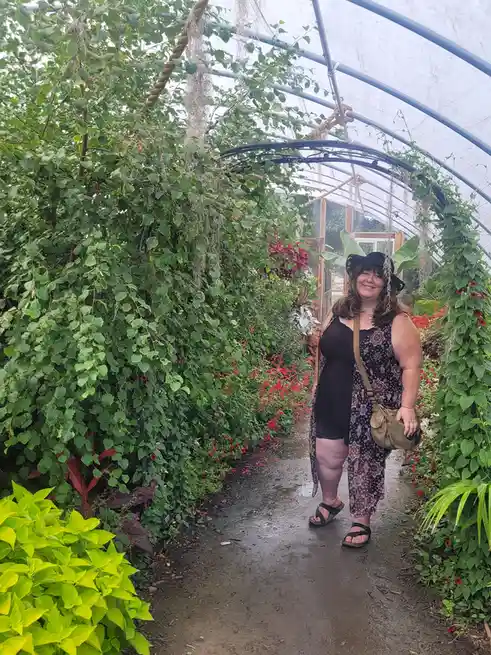 A smiling woman stands in a lush greenhouse pathway, surrounded by dense green foliage and red flowers, wearing a black hat…