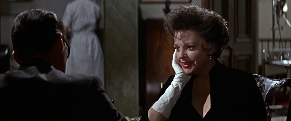 a woman in a black dress and white glove smiles knowingly while looking at a man in a suit, with a nurse visible in the…