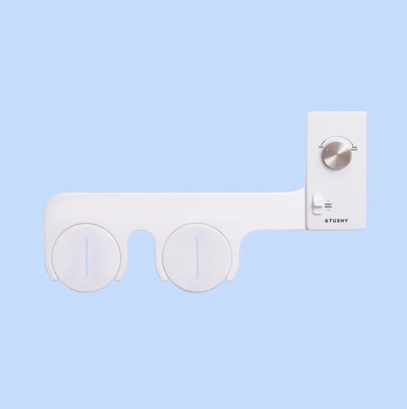A white Tushy bidet sprayer is shown against a light blue background, featuring two circular nozzles and a control panel…