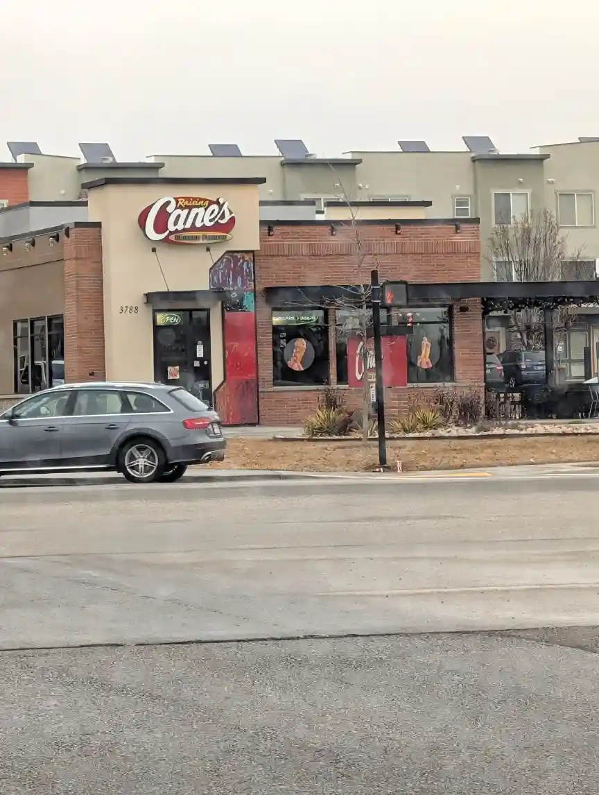 A street-level photo shows a Raising Cane's restaurant with its distinctive red and white sign, brick facade, and windows…